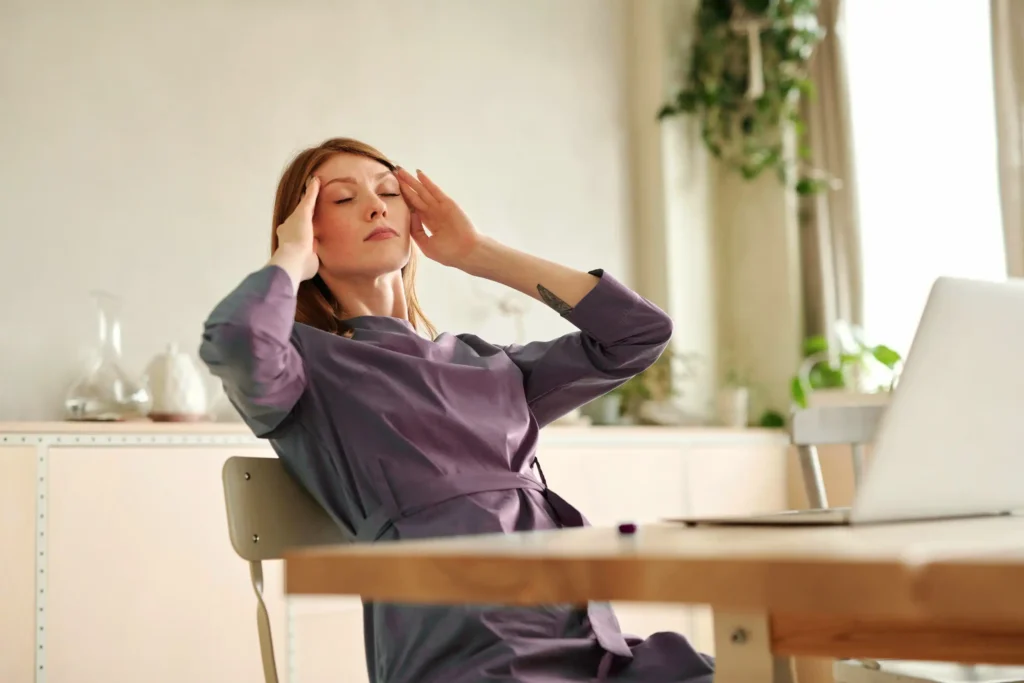 Person at a desk with eyes closed, touching temples beside a laptop, feeling overwhelmed.