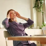 Person at a desk with eyes closed, touching temples beside a laptop, feeling overwhelmed.