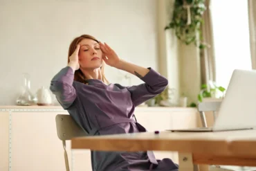 Person at a desk with eyes closed, touching temples beside a laptop, feeling overwhelmed.