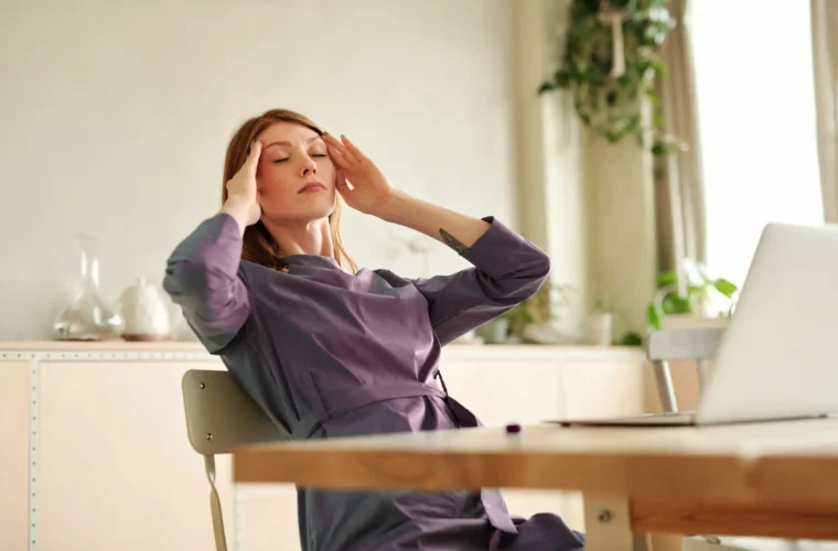 Person at a desk with eyes closed, touching temples beside a laptop, feeling overwhelmed.